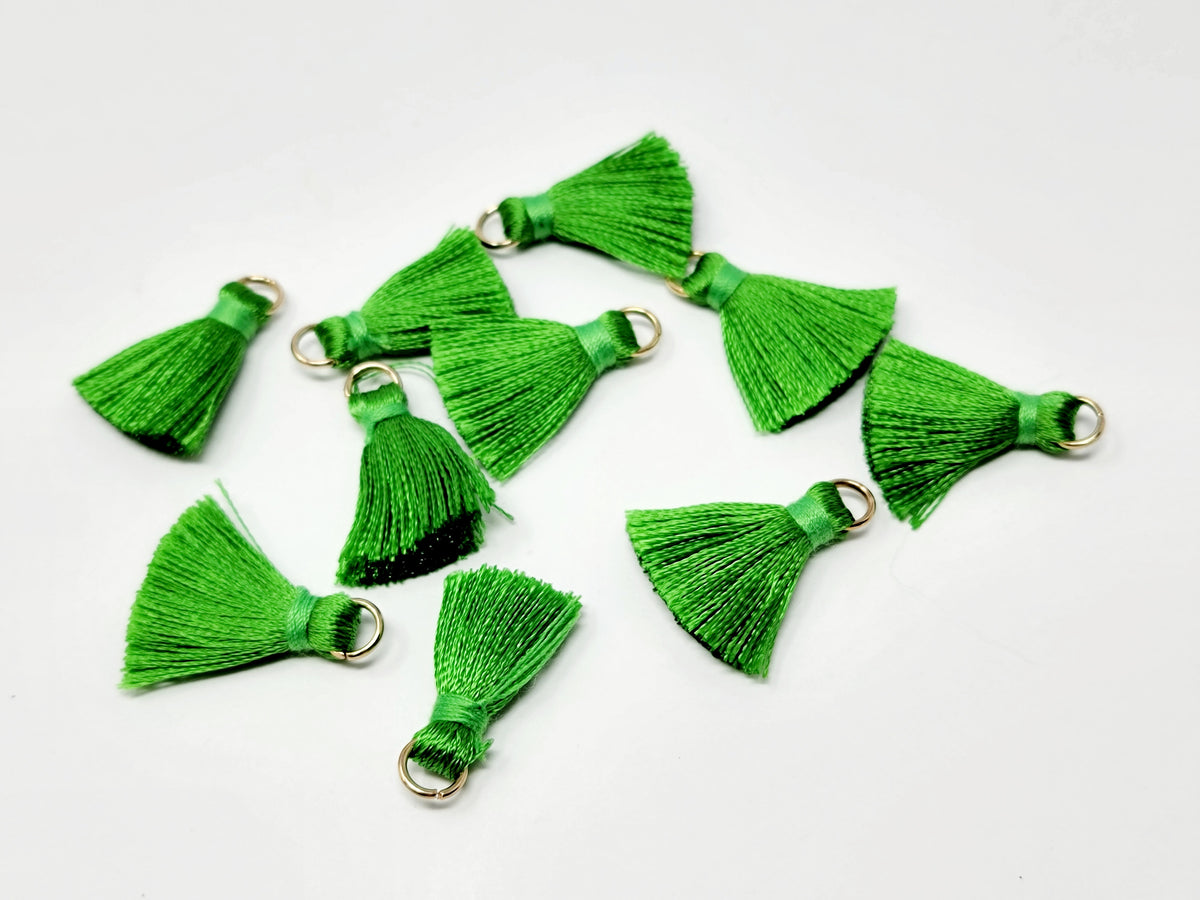 Scattered 2.5cm green cotton tassels with gold-tone jump rings on a white background