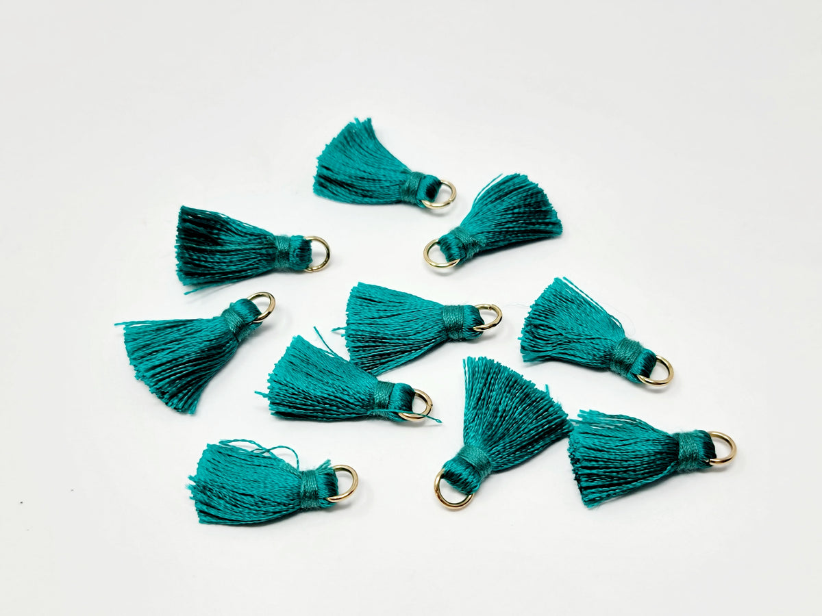 Scattered 2.5cm teal cotton tassels with gold-tone jump rings on a white background
