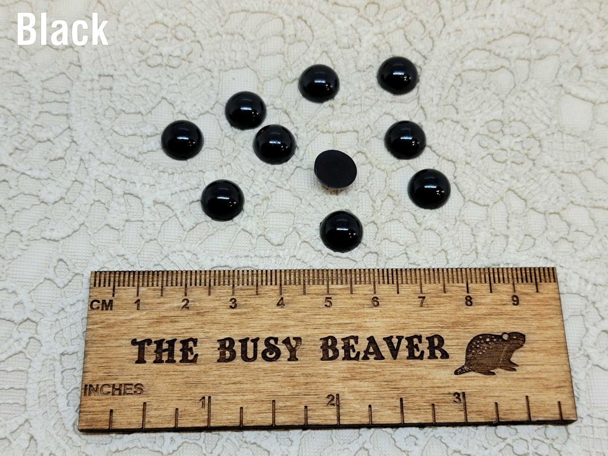 Ten 10mm black pearl-look resin half round cabochons (cabs) beside a ruler