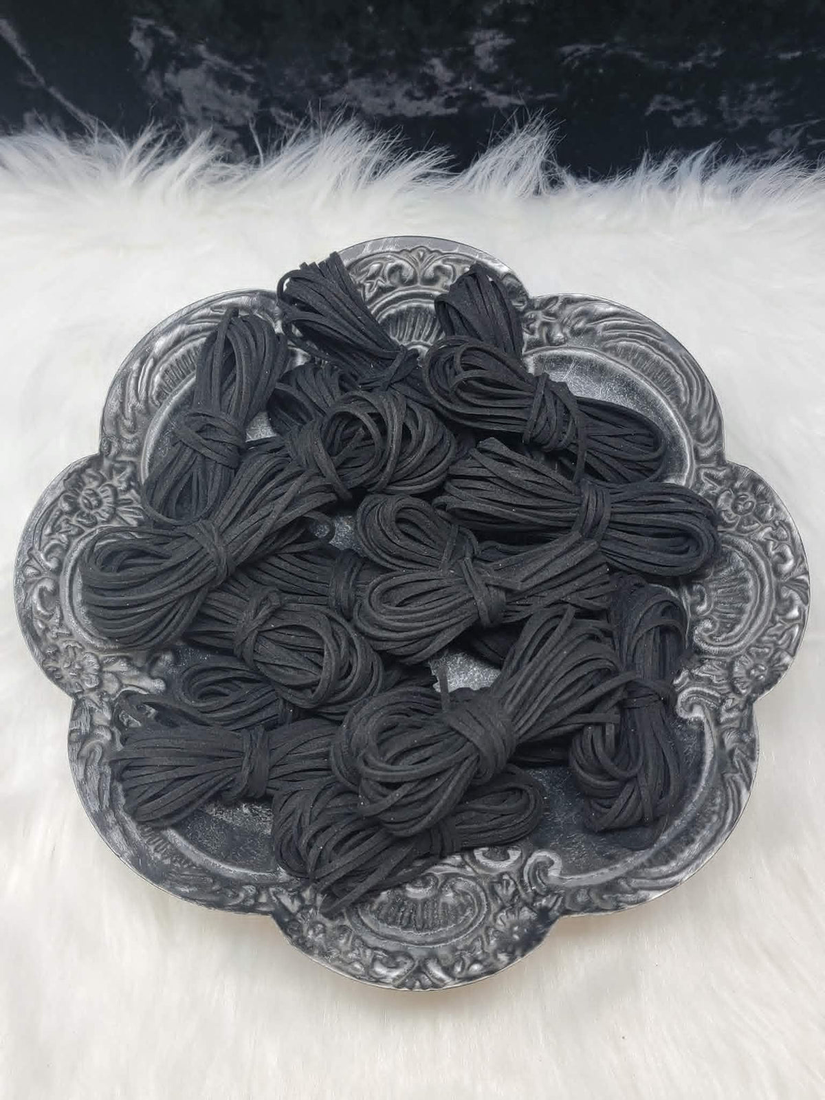 Bundles of 2.7mm black suede leather lacing coiled on a dark scalloped dish