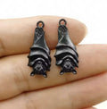 Two dark grey 11 x 27mm hanging bat charms with folded wings, held on a fingertip