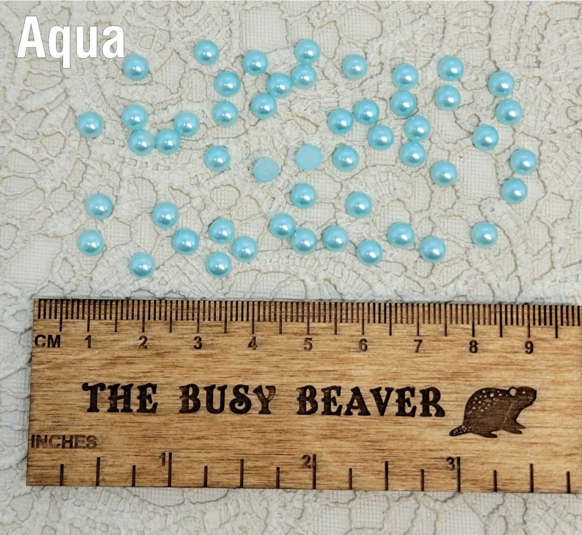 Fifty 5mm aqua pearl-look resin half round cabochons (cabs) scattered beside a ruler