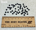 Fifty 5mm black pearl-look resin half round cabochons (cabs) scattered beside a ruler
