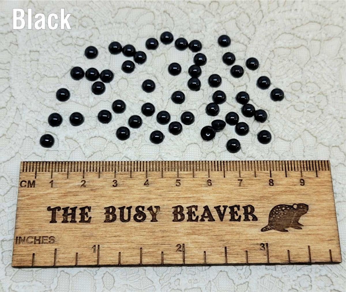 Fifty 5mm black pearl-look resin half round cabochons (cabs) scattered beside a ruler