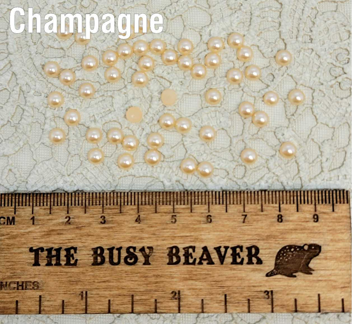Fifty 5mm champagne pearl-look resin half round cabochons (cabs) scattered beside a ruler