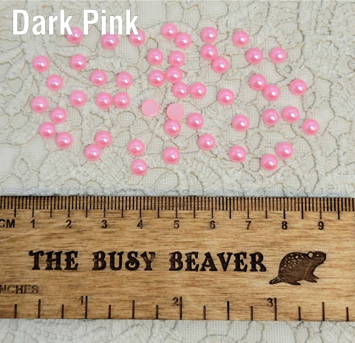 Fifty 5mm dark pink pearl-look resin half round cabochons (cabs) scattered beside a ruler
