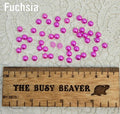 Fifty 5mm fuchsia pearl-look resin half round cabochons (cabs) scattered beside a ruler