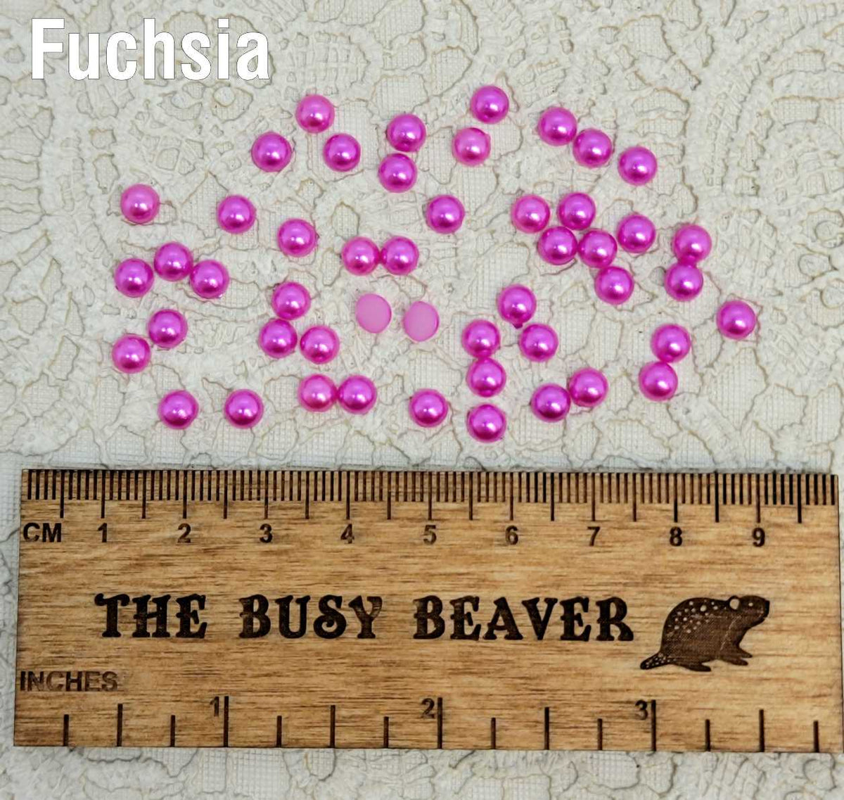 Fifty 5mm fuchsia pearl-look resin half round cabochons (cabs) scattered beside a ruler