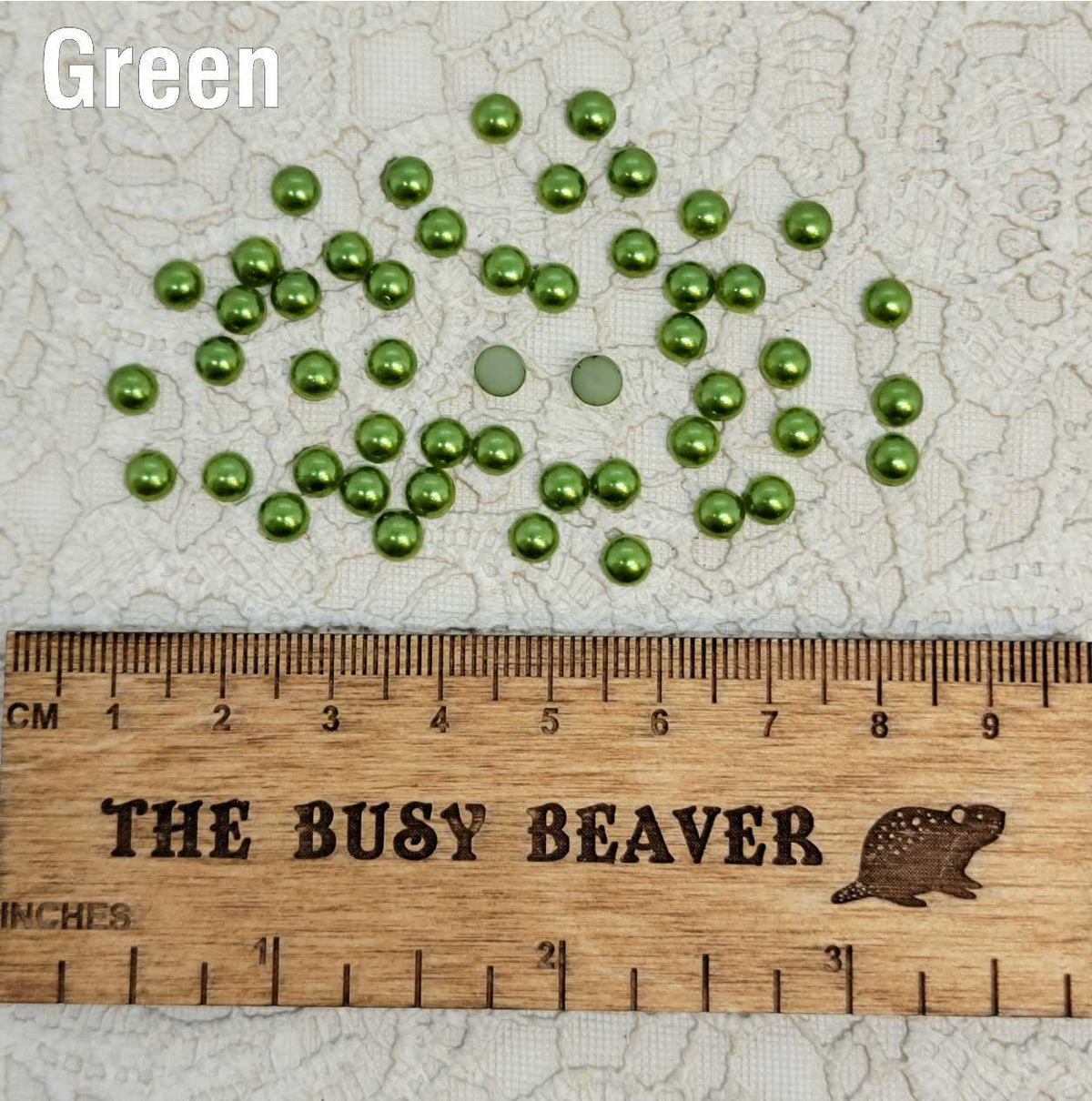 Fifty 5mm green pearl-look resin half round cabochons (cabs) scattered beside a ruler