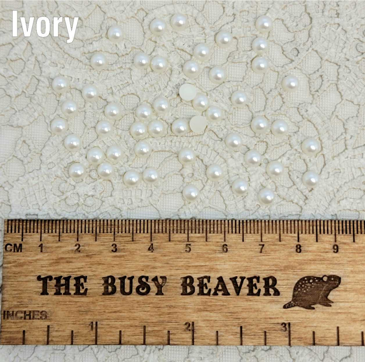 Fifty 5mm ivory pearl-look resin half round cabochons (cabs) scattered beside a ruler