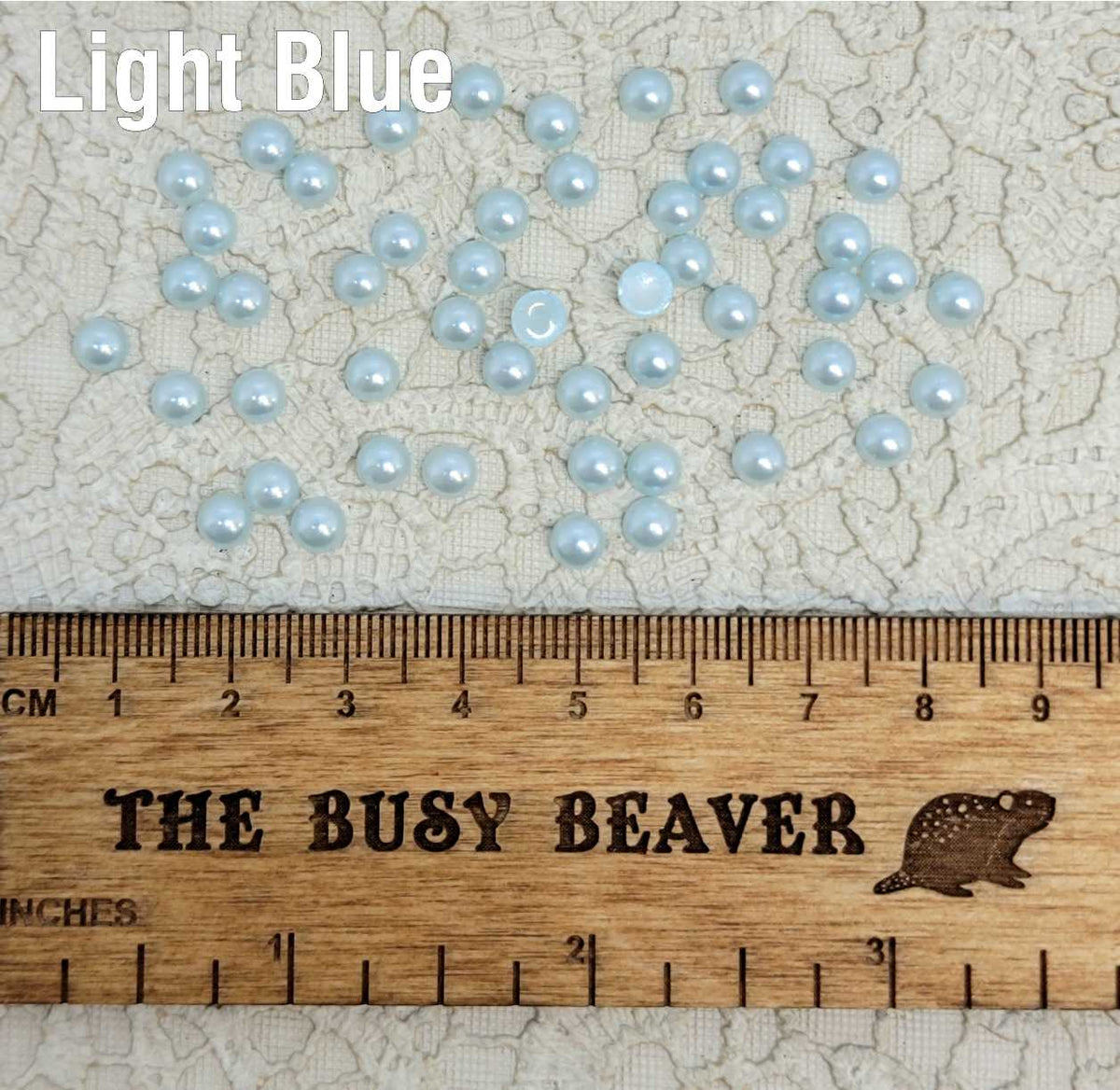Fifty 5mm light blue pearl-look resin half round cabochons (cabs) scattered beside a ruler