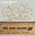 Fifty 5mm light brown pearl-look resin half round cabochons (cabs) scattered beside a ruler