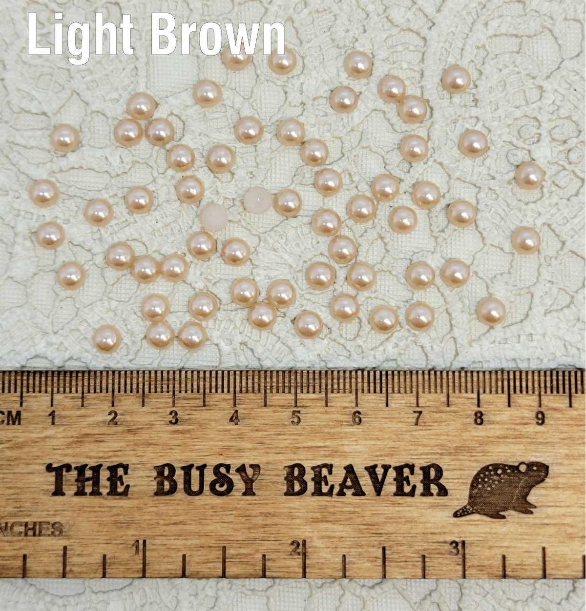 Fifty 5mm light brown pearl-look resin half round cabochons (cabs) scattered beside a ruler