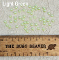 Fifty 5mm light green pearl-look resin half round cabochons (cabs) scattered beside a ruler