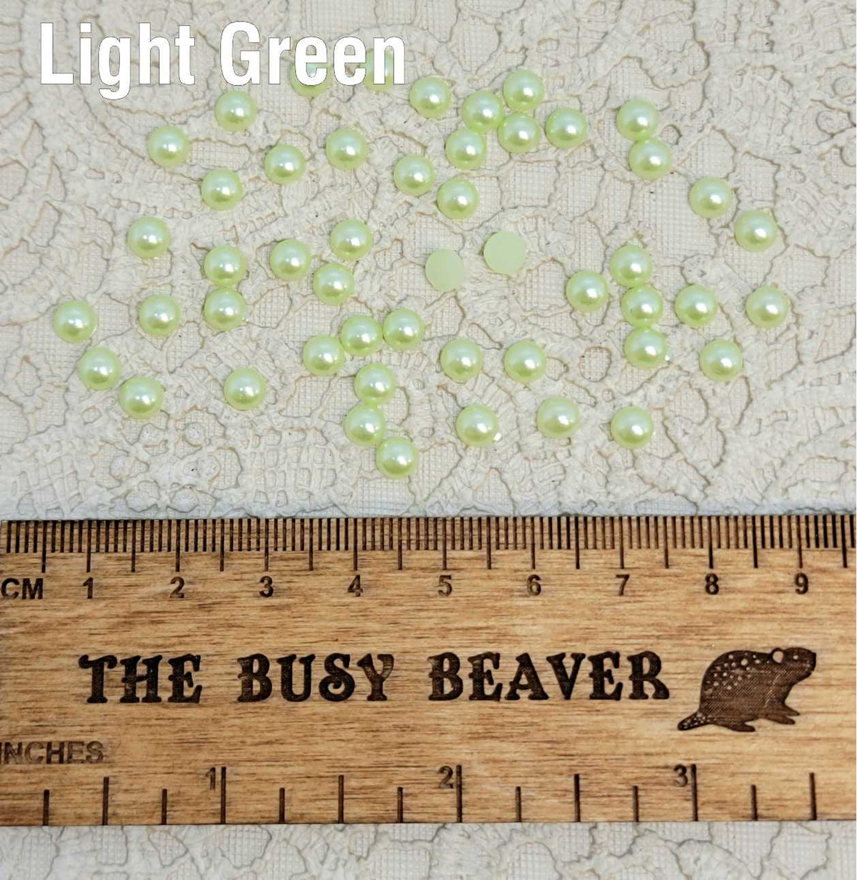 Fifty 5mm light green pearl-look resin half round cabochons (cabs) scattered beside a ruler