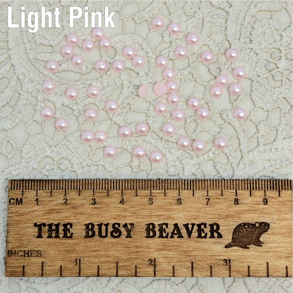 Fifty 5mm light pink pearl-look resin half round cabochons (cabs) scattered beside a ruler