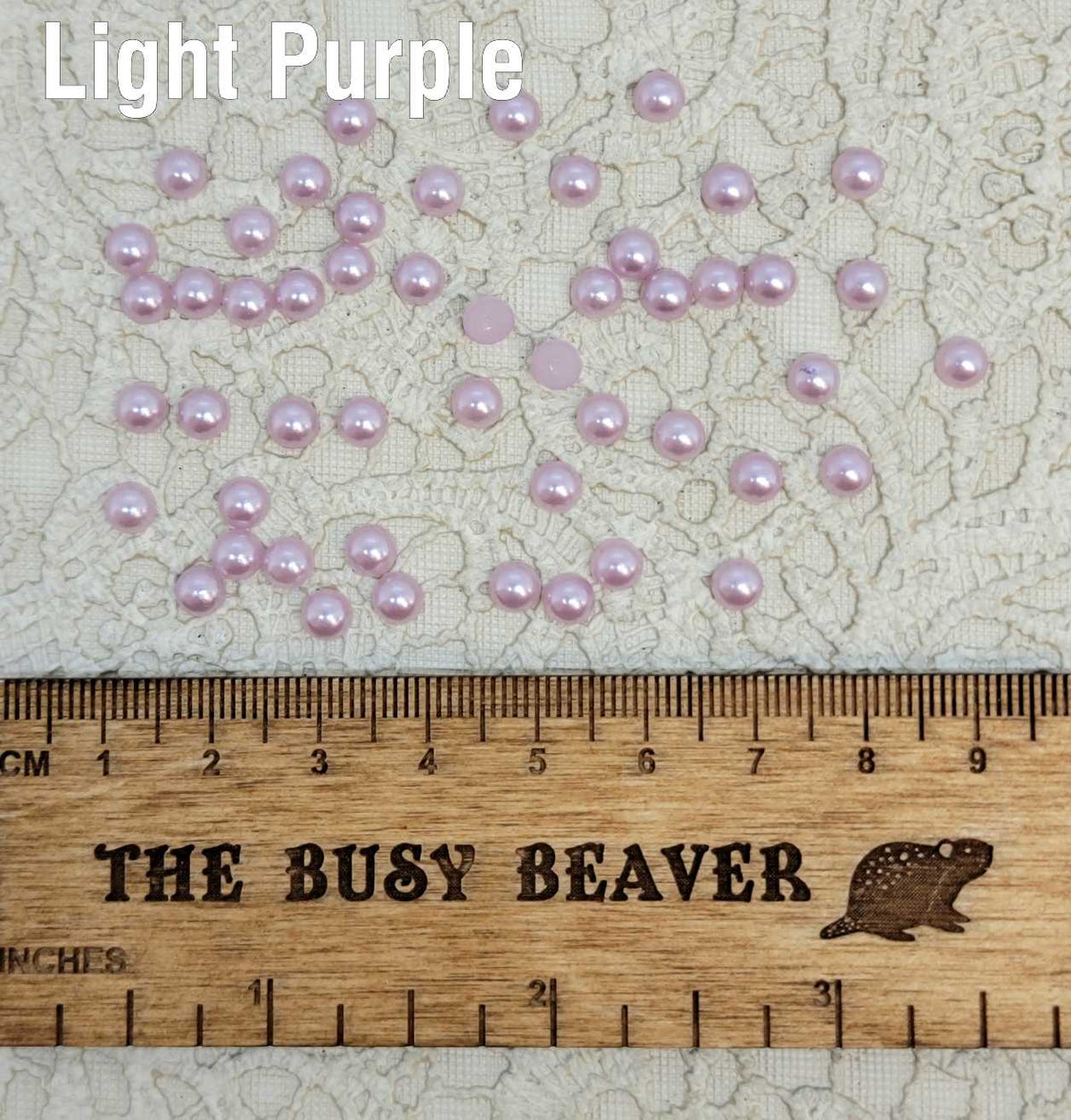 Fifty 5mm light purple pearl-look resin half round cabochons (cabs) scattered beside a ruler