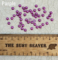 Fifty 5mm purple pearl-look resin half round cabochons (cabs) scattered beside a ruler