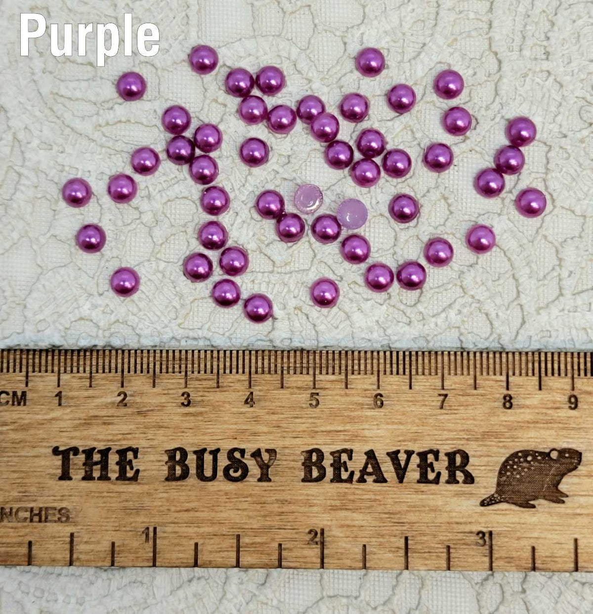 Fifty 5mm purple pearl-look resin half round cabochons (cabs) scattered beside a ruler