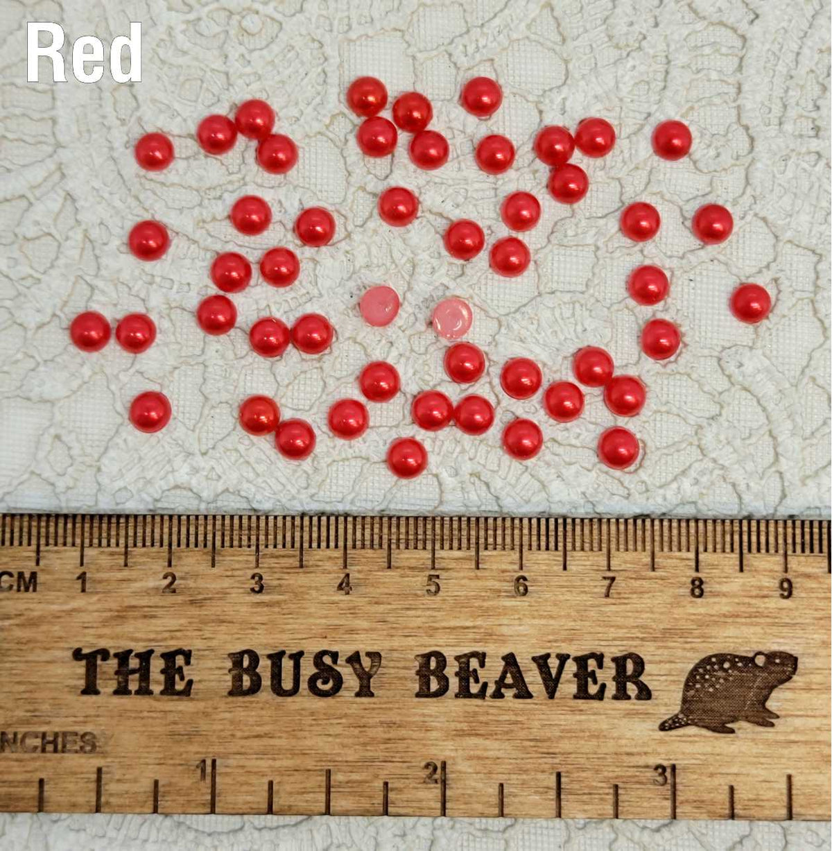 Fifty 5mm red pearl-look resin half round cabochons (cabs) scattered beside a ruler