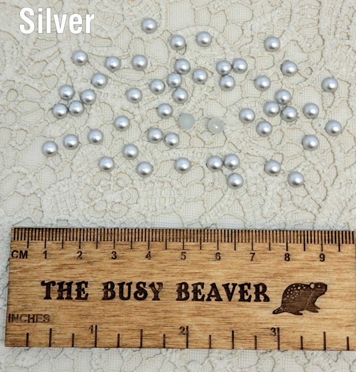 Fifty 5mm silver pearl-look resin half round cabochons (cabs) scattered beside a ruler
