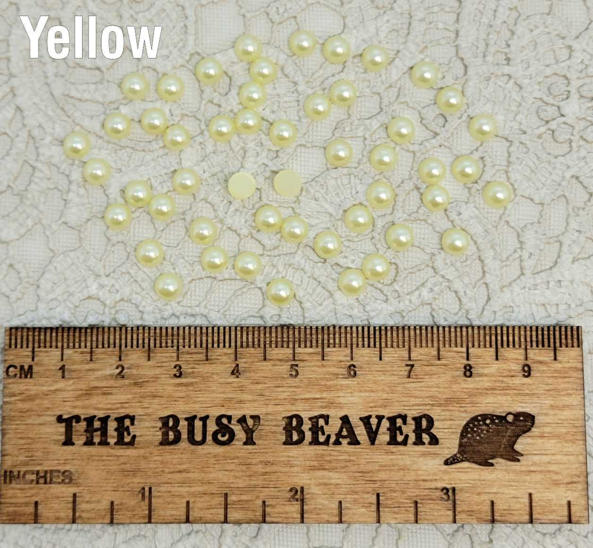 Fifty 5mm yellow pearl-look resin half round cabochons (cabs) scattered beside a ruler